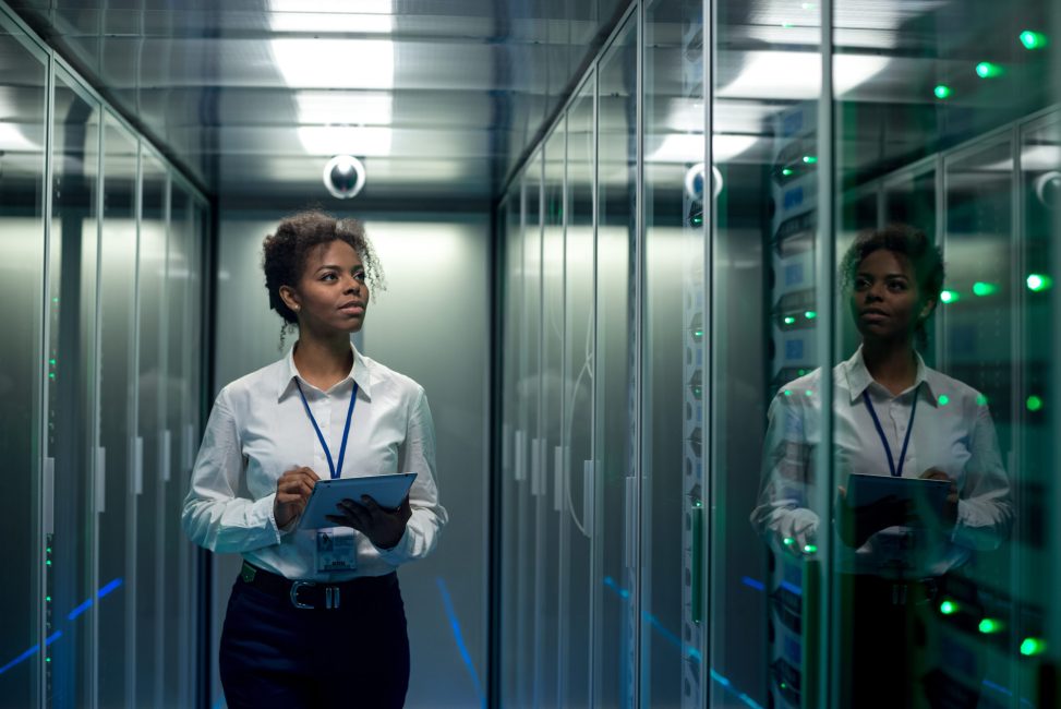 African American woman using tablet while walking in corridor of data center and checking hardware on server racks